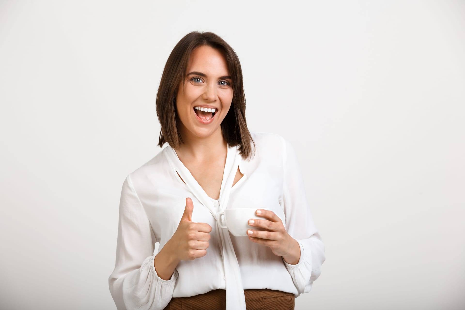 Mulher sorridente de camisa branca faz sinal de positivo segurando uma xícara.