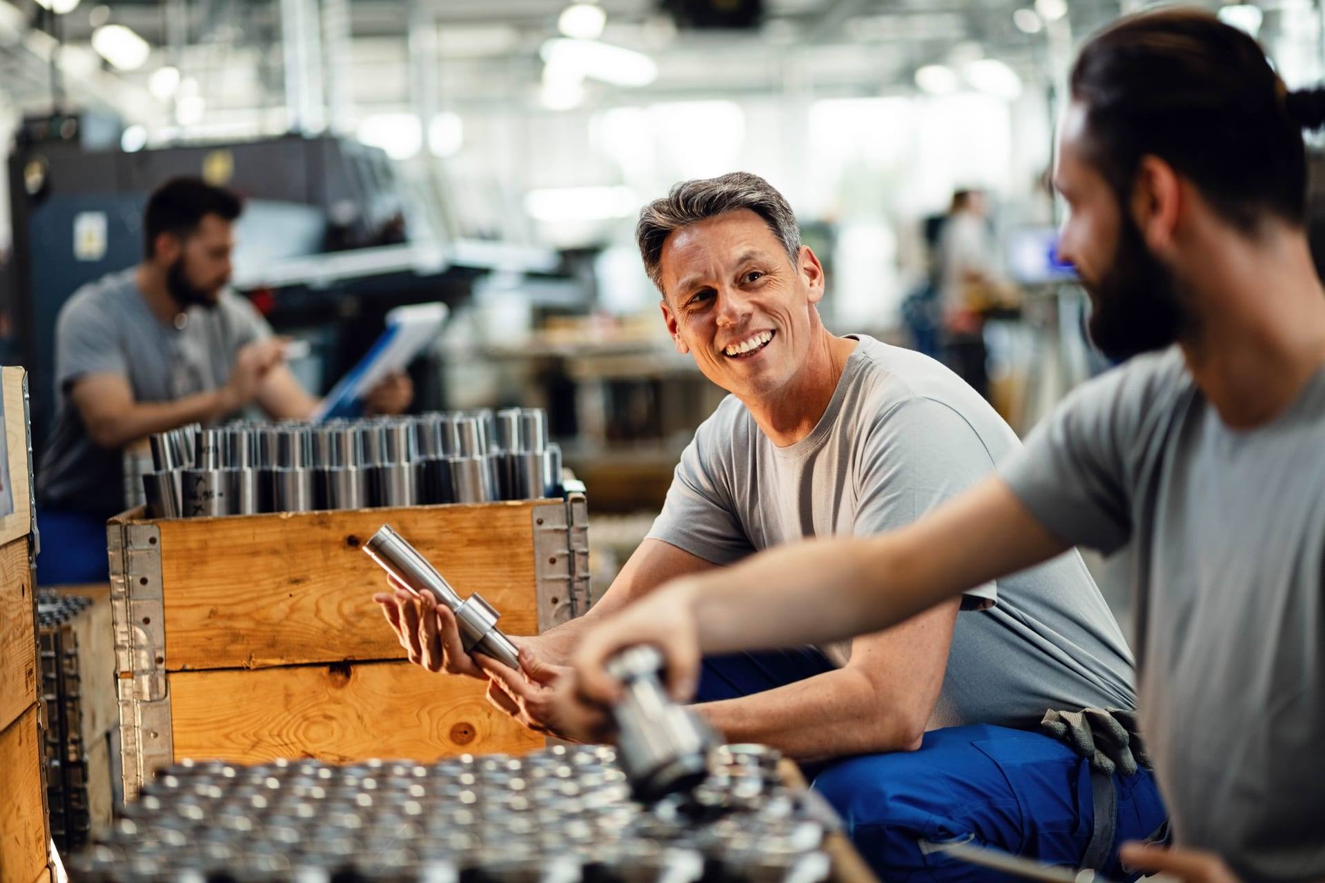 Dois operários conversam sorrindo em uma fábrica de peças metálicas.